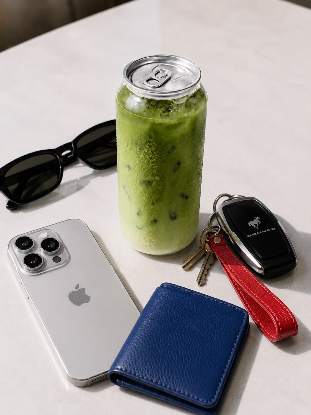 A clear can of iced matcha on a table beside a phone, sunglasses, wallet, and keys